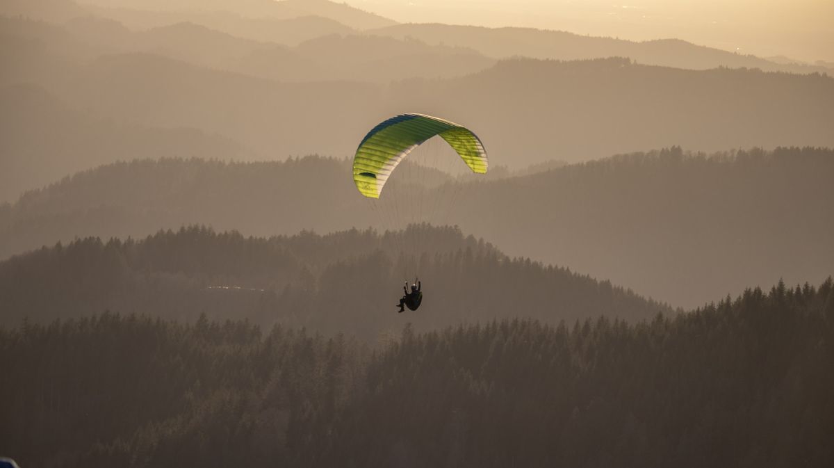 Paraglider with a yellow-green wing over hazy forested ridges